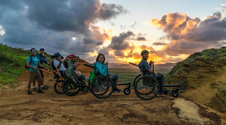 fourpeopleinwheelchairsandtwopeoplestandingontheedgeofamountaintheyaresmilingatthecameraandasunsetisinthebackgroundovermoremountains